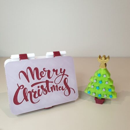 3D‑printed Merry Christmas hinged box beside mini green tree with gold angel and bluegray stars on red stand.