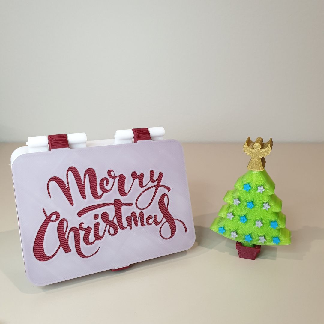 3D‑printed Merry Christmas hinged box beside mini green tree with gold angel and bluegray stars on red stand.